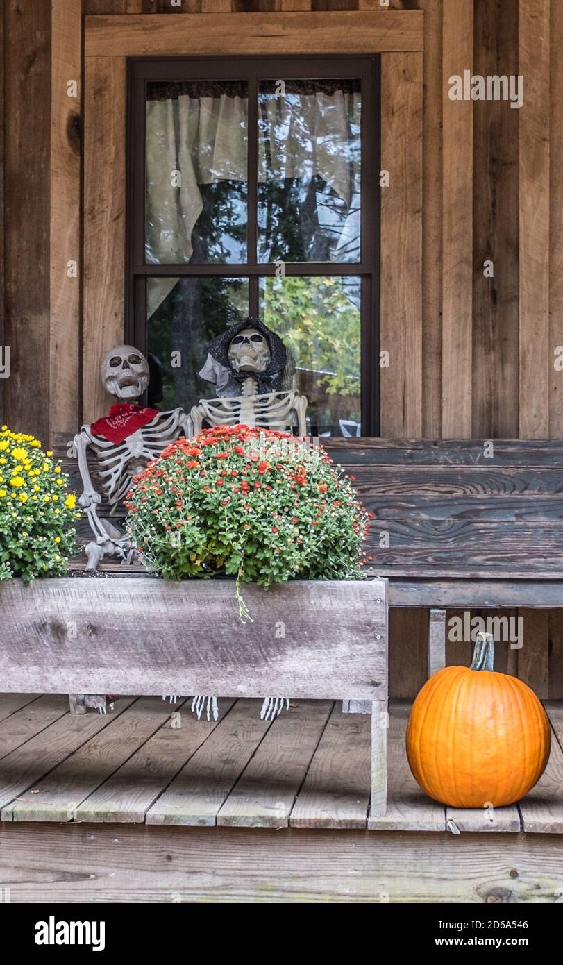 Une paire de squelettes riant assis sur un banc devant une fenêtre sur une véranda à l'avant saluant les gens Entouré d'un décor d'automne pour Halloween Banque D'Images