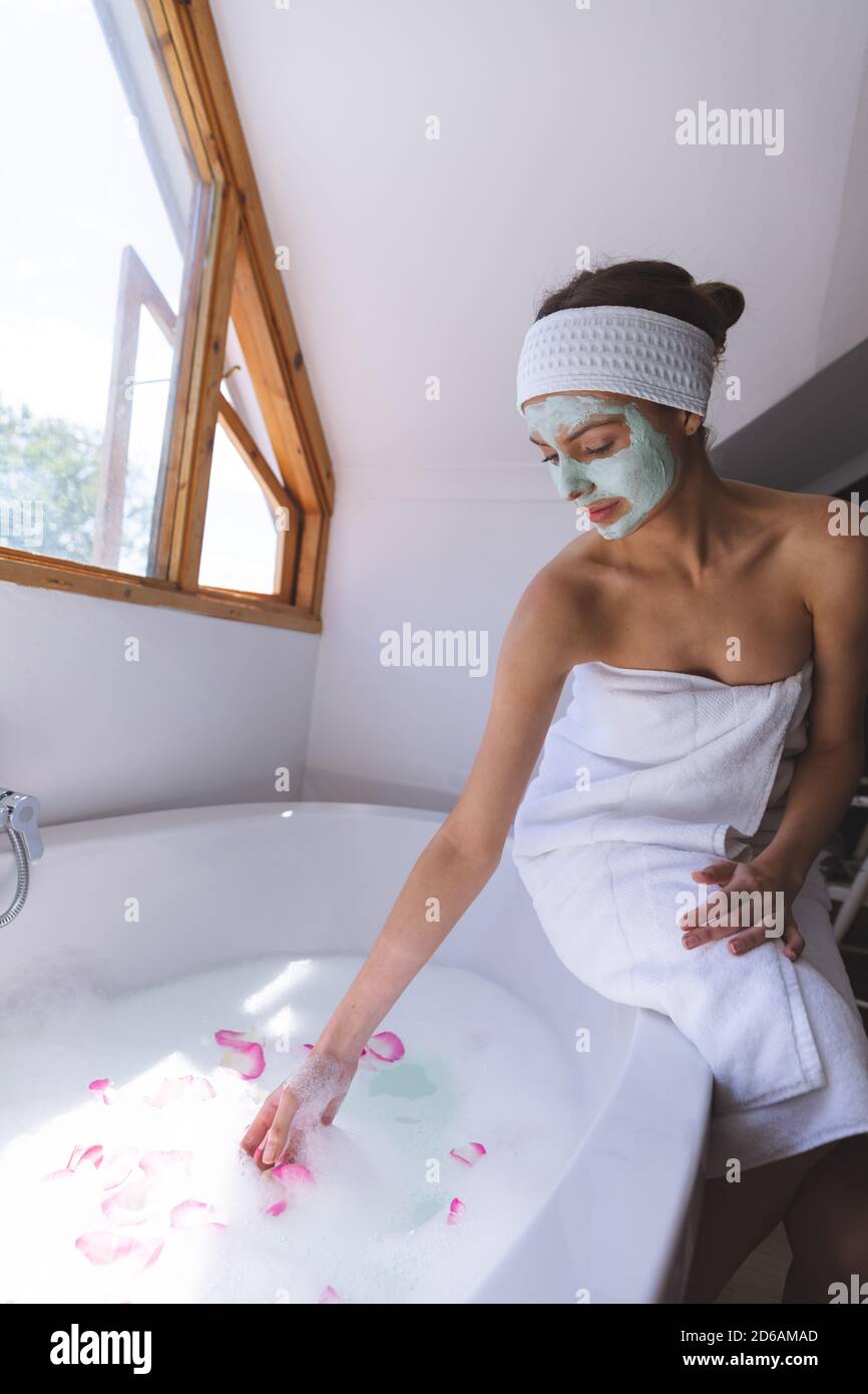 Femme avec masque tombant pétales de rose tout en étant assise bord de la baignoire dans la salle de bains Banque D'Images