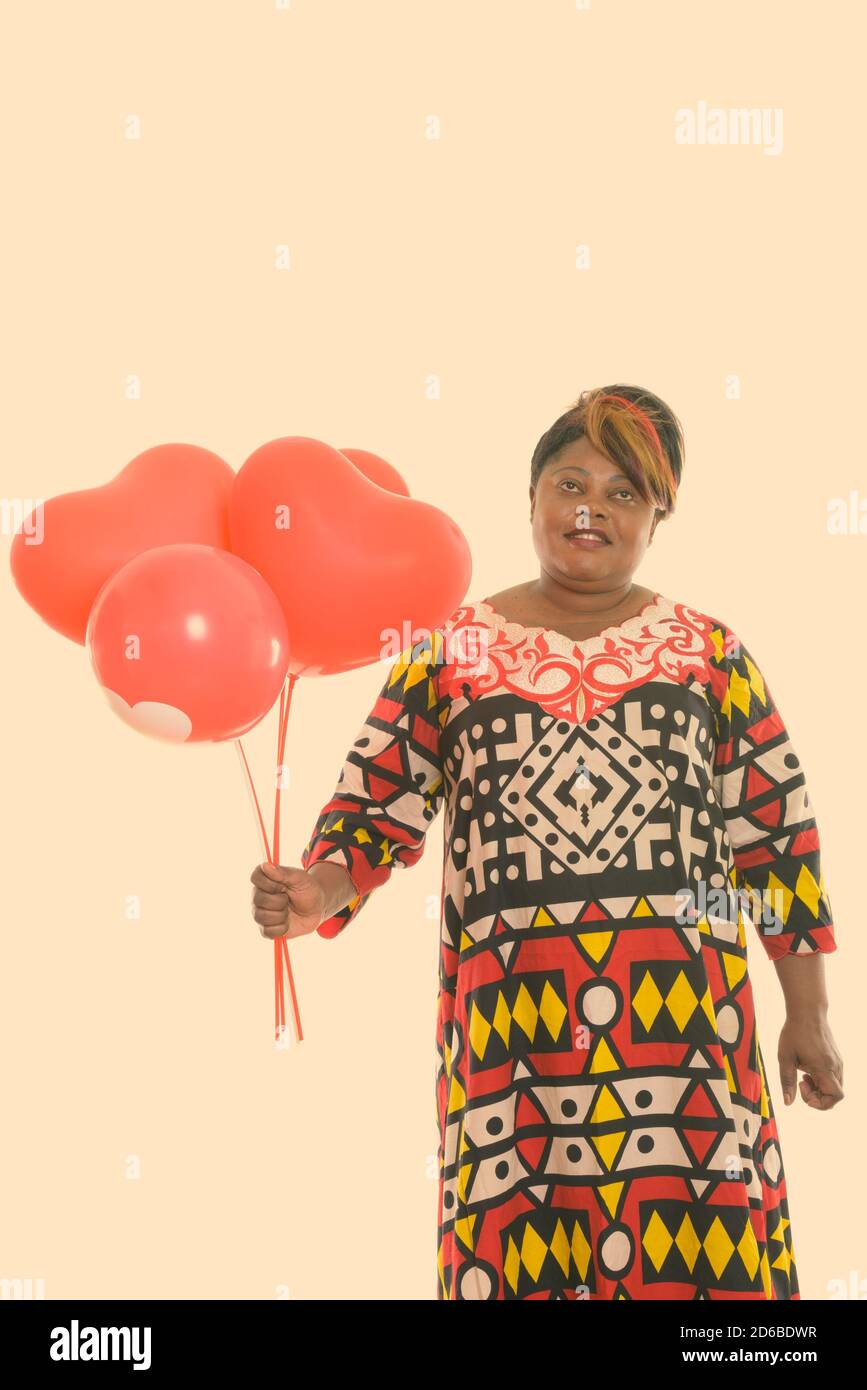 Studio de photo de bonne femme africaine noire graisse souriante et penser tout en tenant un groupe de ballons rouges avec le signe de coeur Banque D'Images