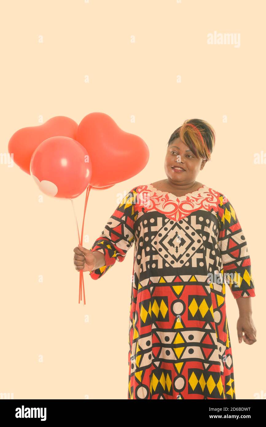 Photo studio d'une femme africaine noire et grasse souriante tenir et regarder un tas de ballons rouges avec le coeur signe Banque D'Images