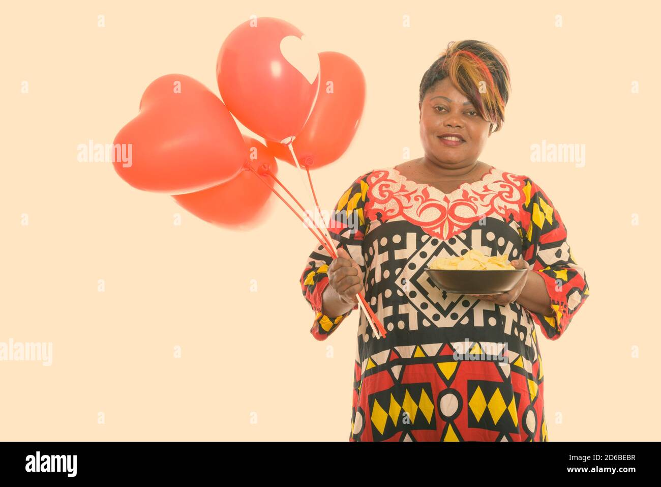 Bonne femme africaine noire grasse souriant tout en tenant un bol de croustilles et bouquet de ballons rouges avec enseigne en forme de cœur Banque D'Images