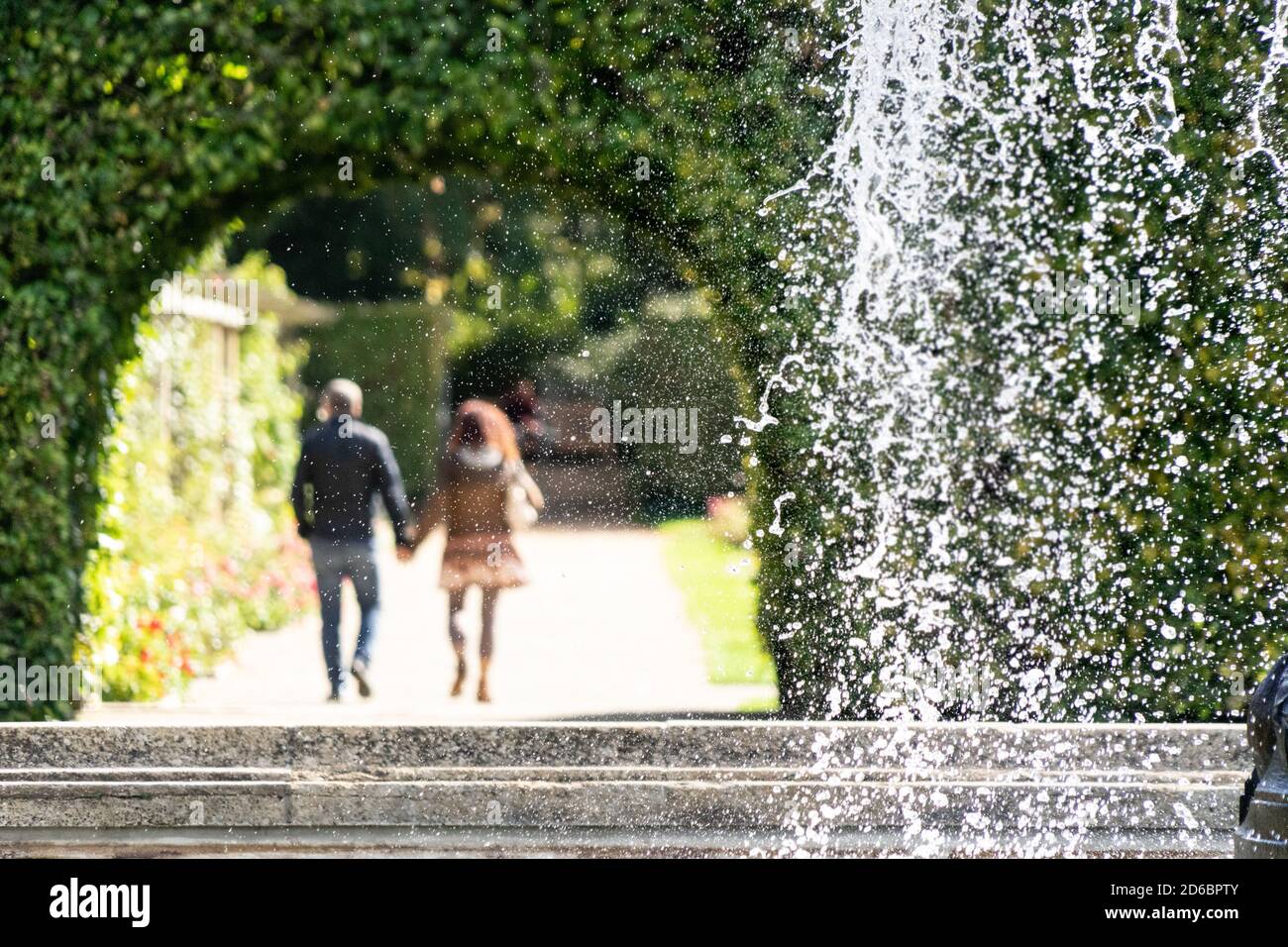 Couple se promenant dans un arrière-plan hors foyer dans un parc ensoleillé avec eau en cascade sur le bord d'un fontaine en premier plan Banque D'Images