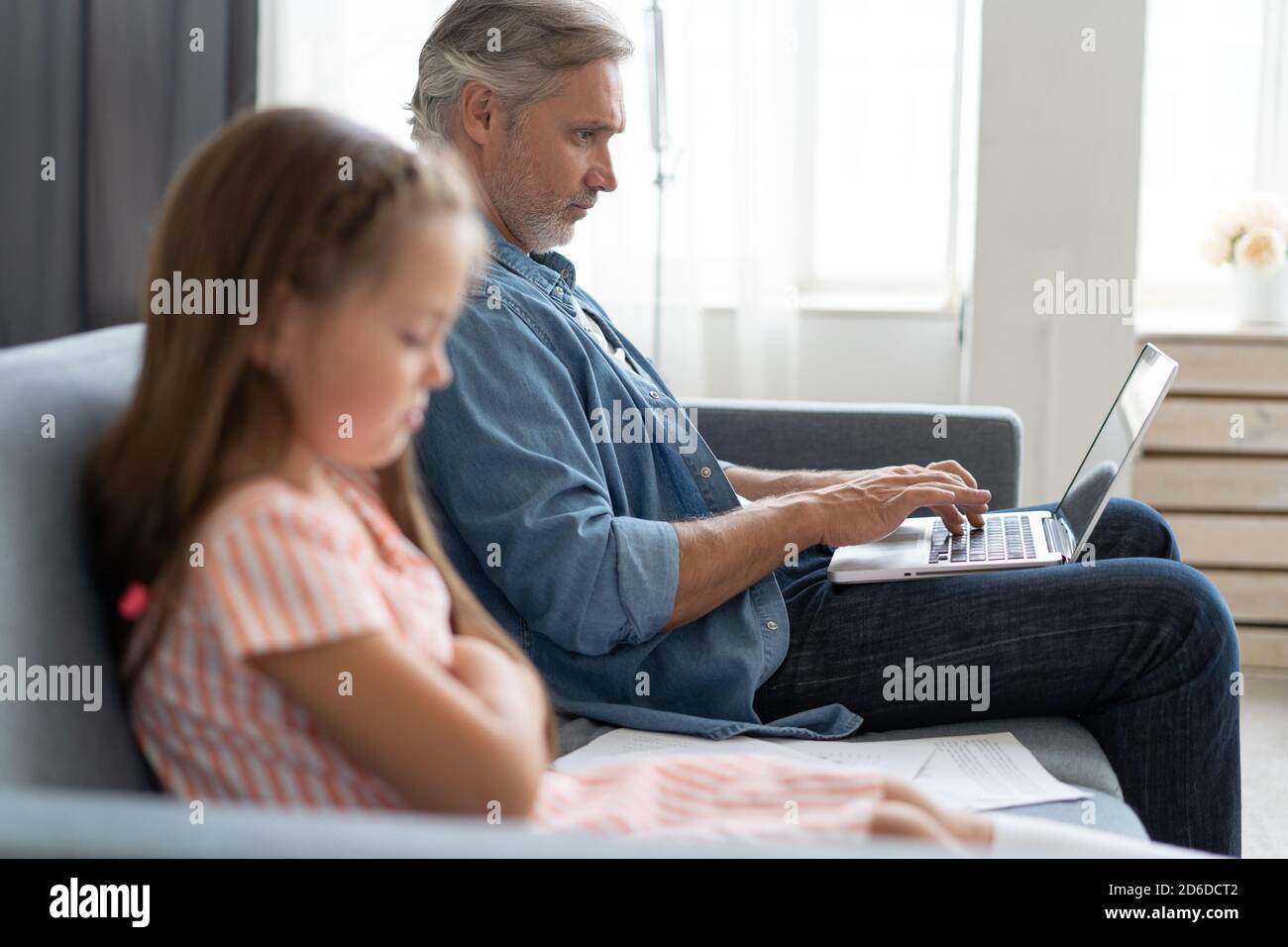Pas de temps pour l'enfant. Papa aux cheveux gris occupé avec un ordinateur portable, travaillant en ligne à la maison, triste ennuyé fille offended assis à proximité Banque D'Images