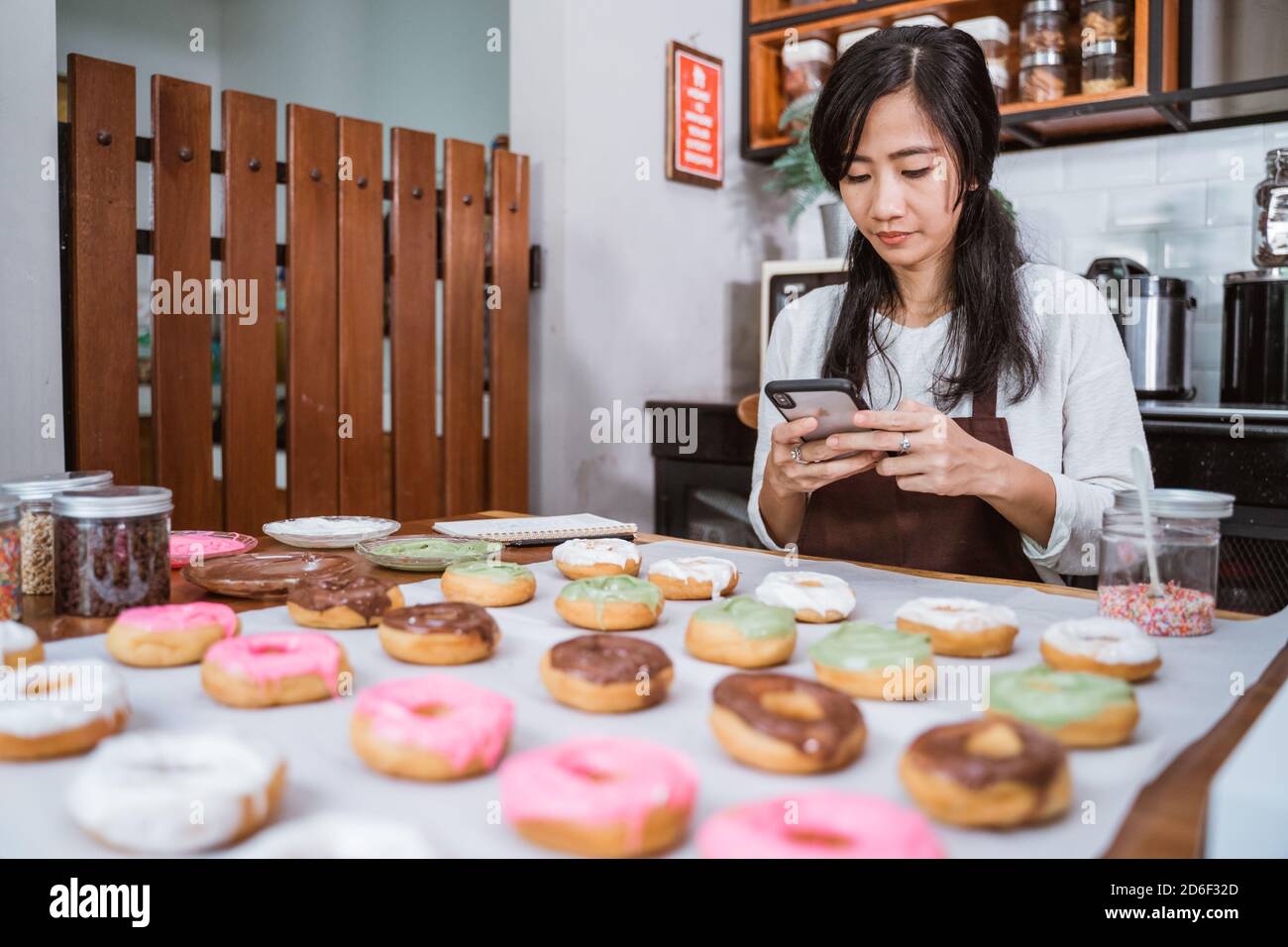 une jeune femme portant un tablier à l'aide d'un téléphone cellulaire après décoration de divers beignets dans la cuisine Banque D'Images