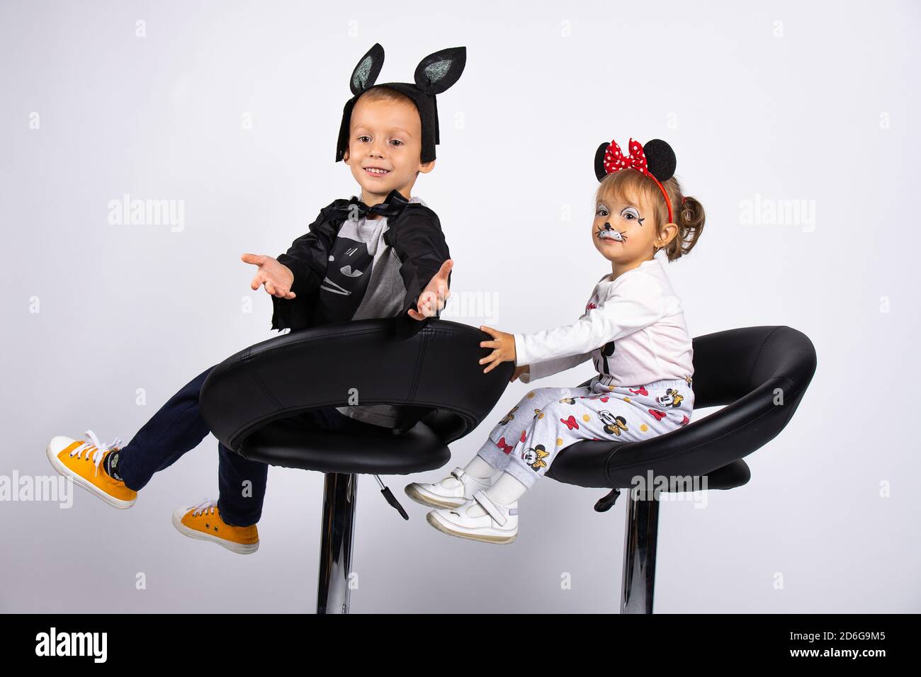Des enfants heureux attendent les vacances. Drôle de garçon et de fille en costumes de carnaval assis dans des chaises noires à l'intérieur sur un fond blanc. Banque D'Images