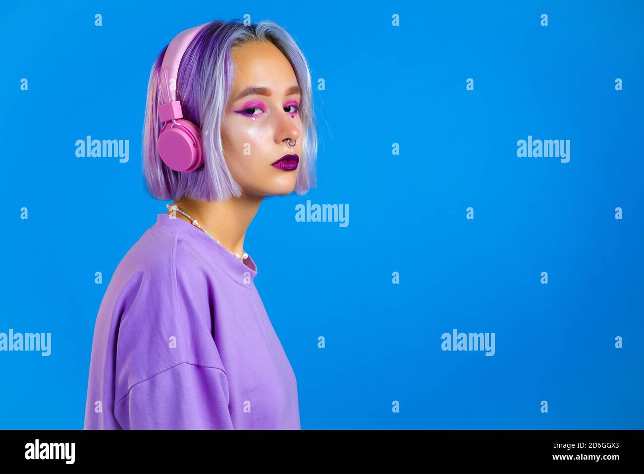 Jolie fille aux cheveux violets teints écoutant de la musique, souriant, dansant dans un casque en studio sur fond bleu. Musique, danse, concept radio Banque D'Images