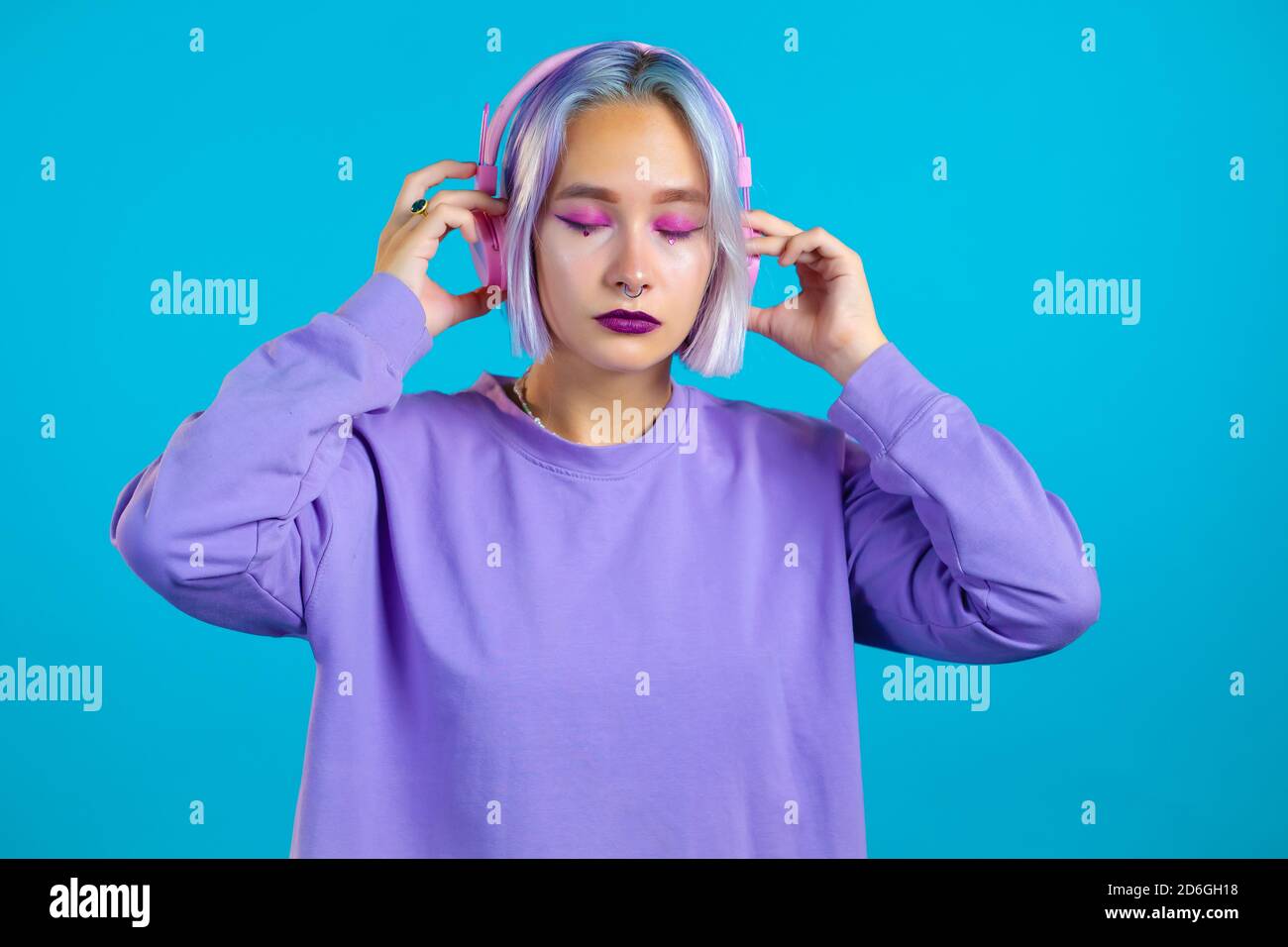 Jolie fille aux cheveux violets teints écoutant de la musique, souriant, dansant dans un casque en studio sur fond bleu. Musique, danse, concept radio Banque D'Images