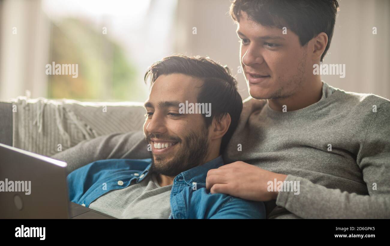 Le couple Sweet Male Queer passe du temps à la maison. Ils sont couchés sur un canapé et utilisent l'ordinateur portable. Ils naviguer en ligne. La main du partenaire est autour de son Lover Banque D'Images