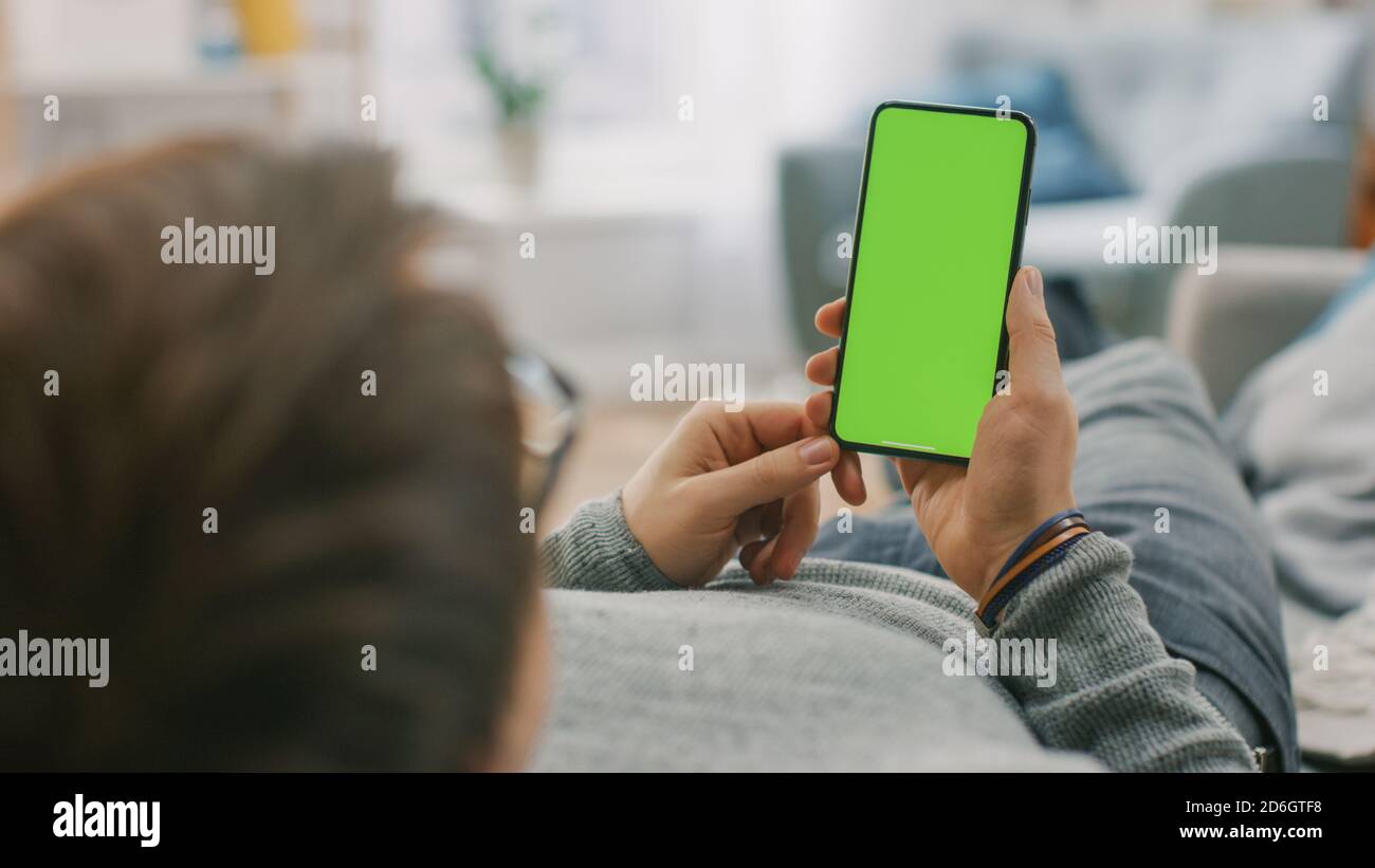 Homme à la maison allongé sur un canapé à l'aide d'un smartphone avec écran vert Mock-up, gestes de défilement. Guy utilisant le téléphone mobile, Internet social Networks Browsing Banque D'Images