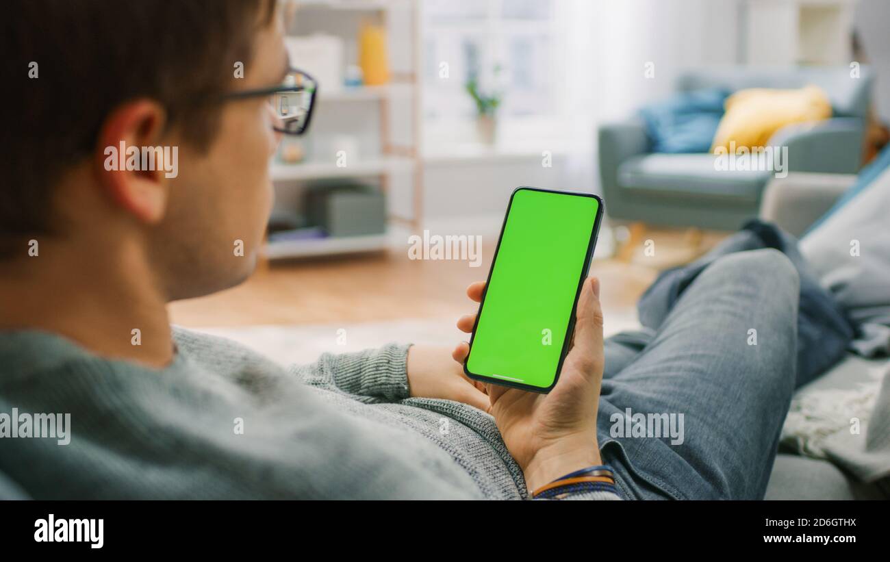 Homme à la maison reposant sur un canapé à l'aide d'un smartphone avec écran de maquette vert. Guy utilisant le téléphone mobile, Internet social Networks Browsing. Banque D'Images
