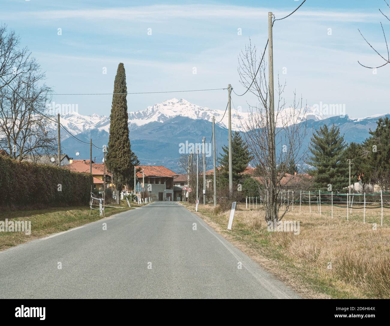 La route de campagne entre dans une zone bâtie avec des montagnes enneigées en arrière-plan. Piémont, Italie Banque D'Images