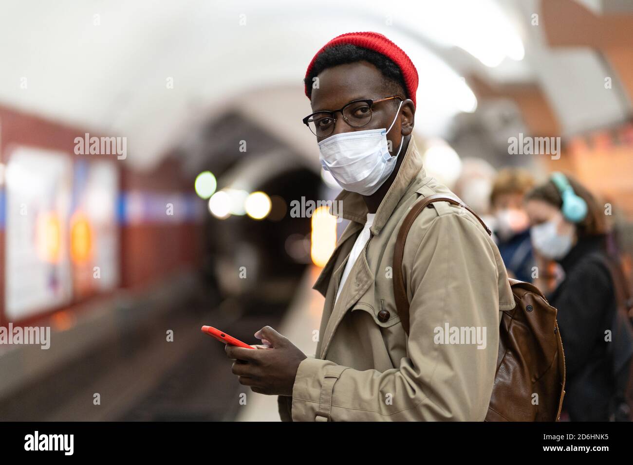 Homme millénaire afro-américain en trench coat, chapeau rouge portant un masque de protection contre le covid-19, virus de la grippe, attendant le train au métro stat Banque D'Images