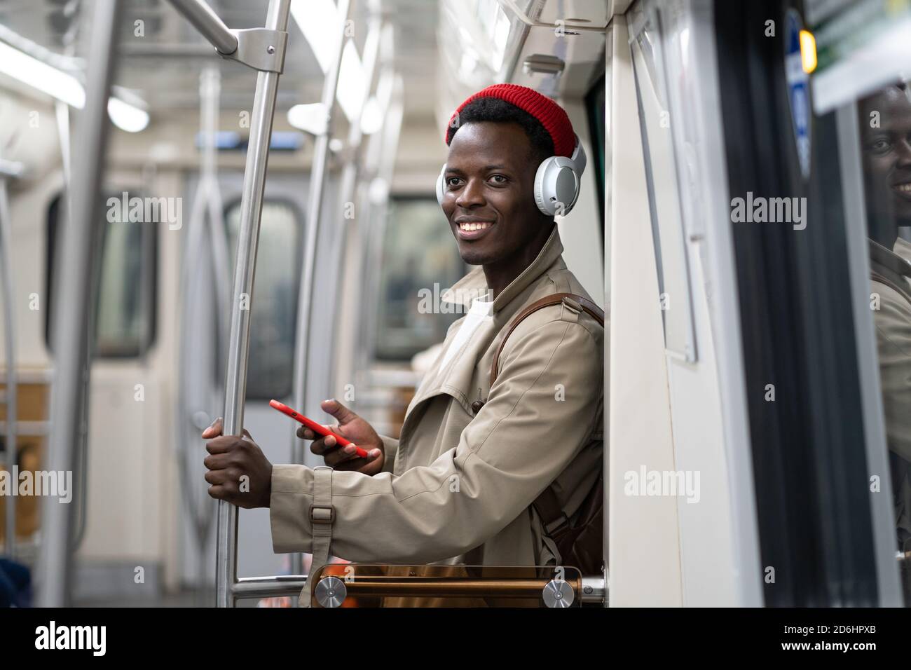 Joyeux homme millénaire afro-américain en chapeau rouge, tranch coat stand dans le métro, en utilisant le téléphone mobile, écoute de la musique avec des écouteurs sans fil dedans Banque D'Images
