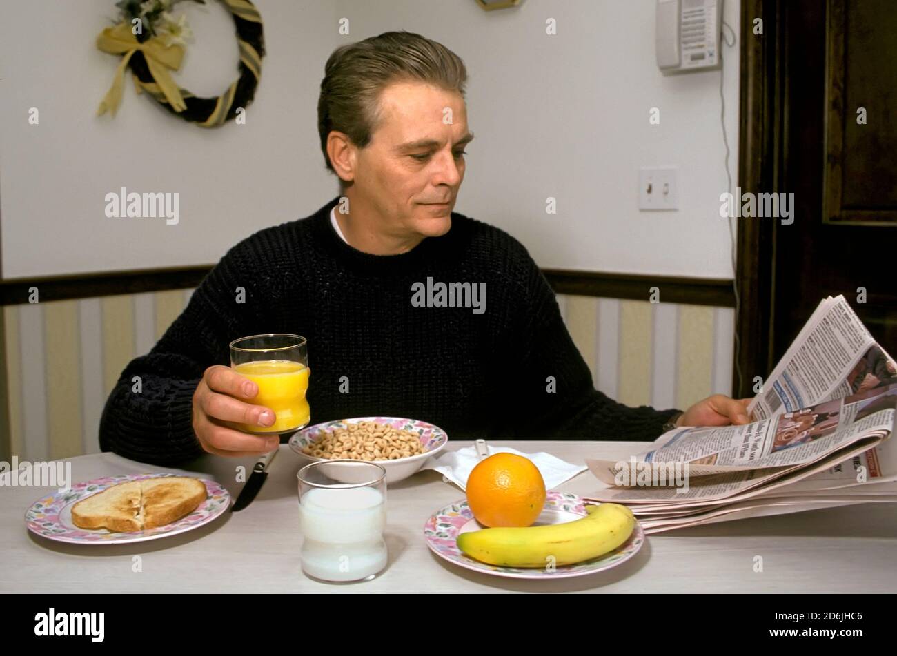 L'homme adulte mange un petit-déjeuner sain comprenant des fruits, des céréales entières, du jus et du lait à faible teneur en matières grasses lors de la lecture du journal Banque D'Images