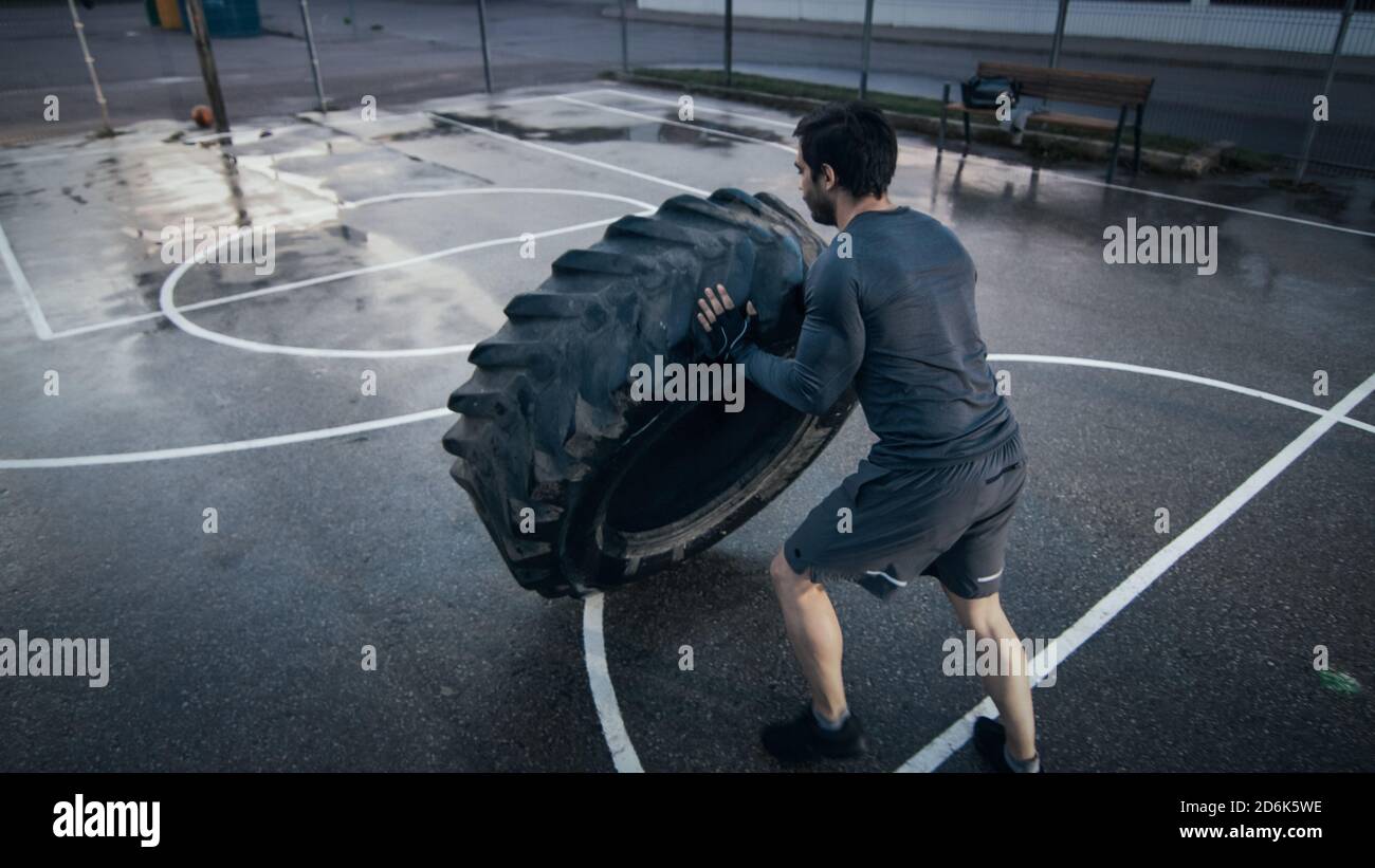 Un jeune homme à la coupe musclée et des gants de sport font des exercices sur un terrain de basket-ball extérieur clôturé. Il bascule un gros pneu lourd dans un Banque D'Images