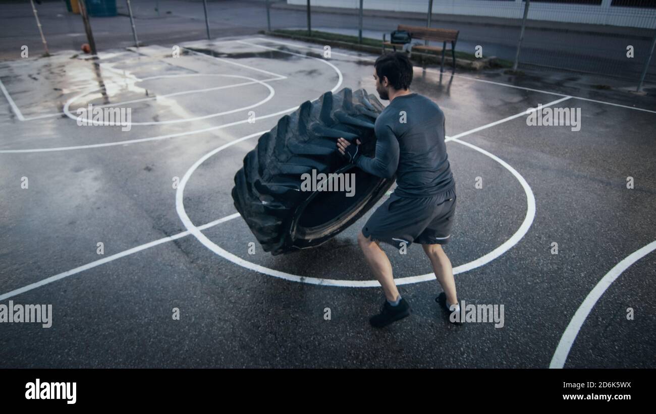 Un jeune homme à la coupe musclée et des gants de sport font des exercices sur un terrain de basket-ball extérieur clôturé. Il bascule un gros pneu lourd dans un Banque D'Images