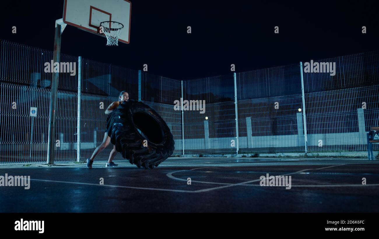 Belle forme physique énergique fille fait des exercices dans un terrain de basket-ball extérieur clôturé. Elle bascule un gros pneu lourd dans une nuit brumeuse après la pluie Banque D'Images