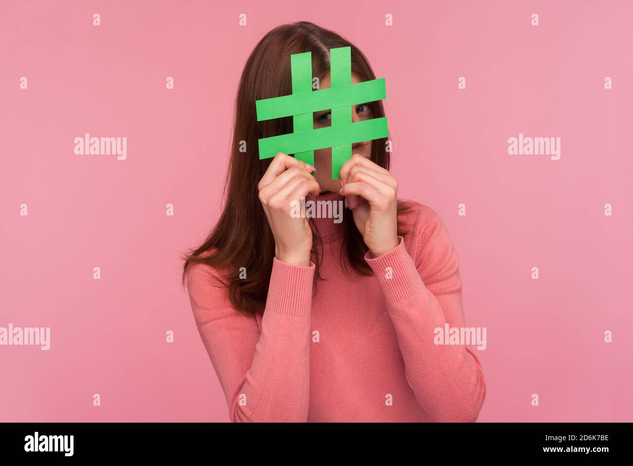 Curieuse brune femme en chandail rose regardant à travers le signe vert hashtag, à la recherche de bons postes dans les médias sociaux, espionnage. Prise de vue en studio en intérieur Isola Banque D'Images