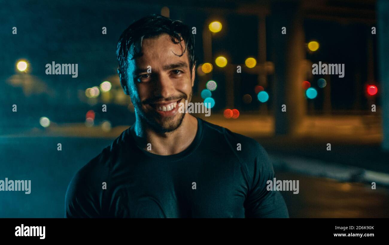 Photo portrait d'un jeune homme athlétique en tenue de sport sombre donnant un sourire après un entraînement de rue lors d'une journée de pluie. Il est dans une soirée urbaine Banque D'Images