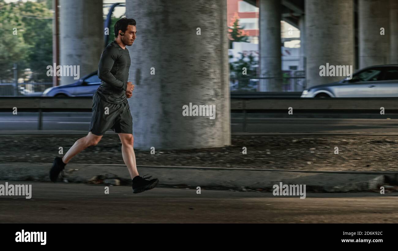 Athletic Young Man dans les écouteurs et les tenues de sport est le jogging dans la rue. Il court dans un environnement urbain sous un pont avec Cars dans le Banque D'Images