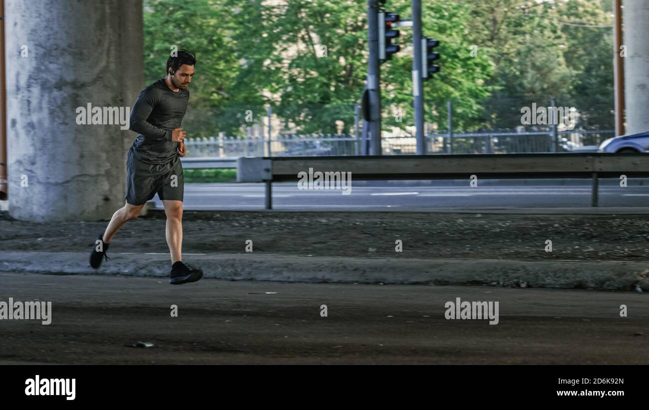 Athletic Young Man dans les écouteurs et les tenues de sport est le jogging dans la rue. Il court dans un environnement urbain sous un pont avec Cars dans le Banque D'Images