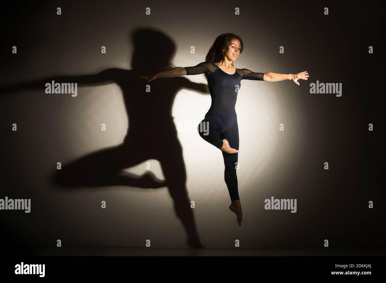 la femme dans l'obscurité effectue le saut de gymnastique, sur un fond blanc il y a une ombre d'une forme. Photographie en studio. Banque D'Images