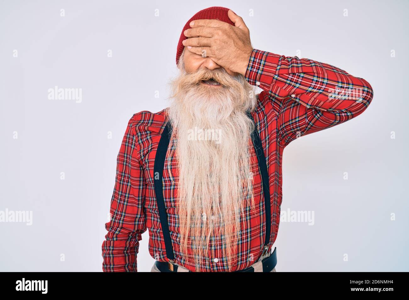 Homme âgé avec cheveux gris et longue barbe portant un look hipster avec  casquette de laine souriant et riant avec la main sur le visage couvrant  les yeux pour la surprise. bli