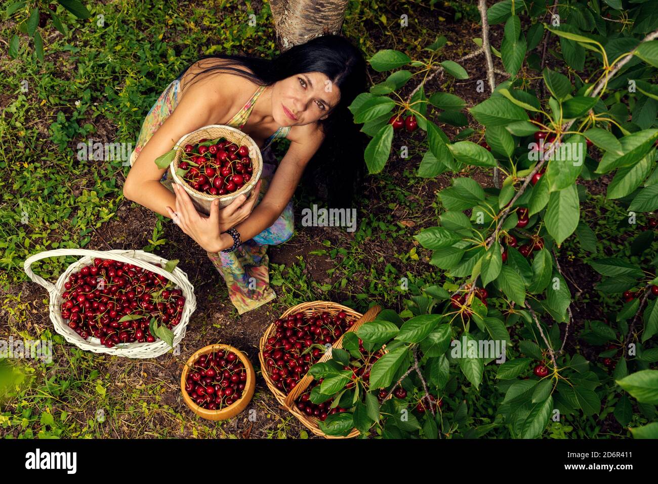 Belle jeune femme portant une robe colorée sous le cerisier avec deux paniers en osier et deux seaux pleins de mûres Fruits foyer sélectif Banque D'Images