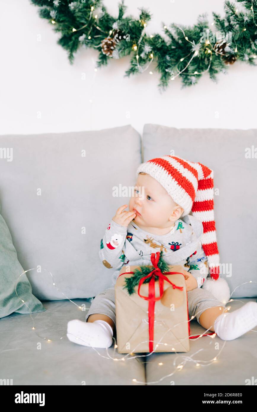 Le petit garçon couvant est assis dans un chapeau de père Noël et un costume de vacances sur un canapé gris avec un grand cadeau avec ruban rouge, contre Mur blanc avec couronne de sapin naturel.CH Banque D'Images