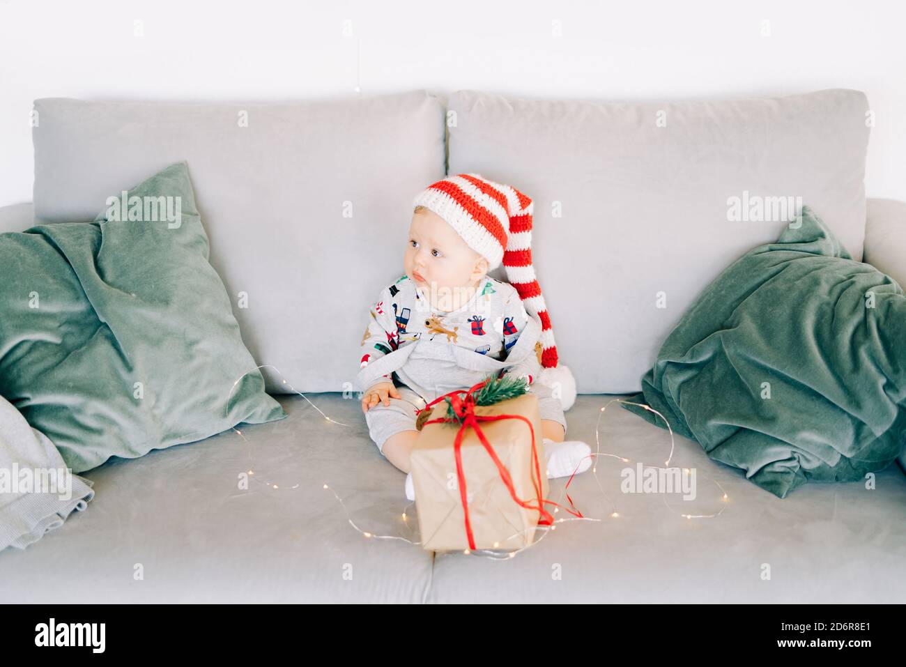 Le petit garçon couvant est assis dans un chapeau de père Noël et un costume de vacances sur un canapé gris avec un grand cadeau avec ruban rouge, contre Mur blanc avec couronne de sapin naturel.CH Banque D'Images