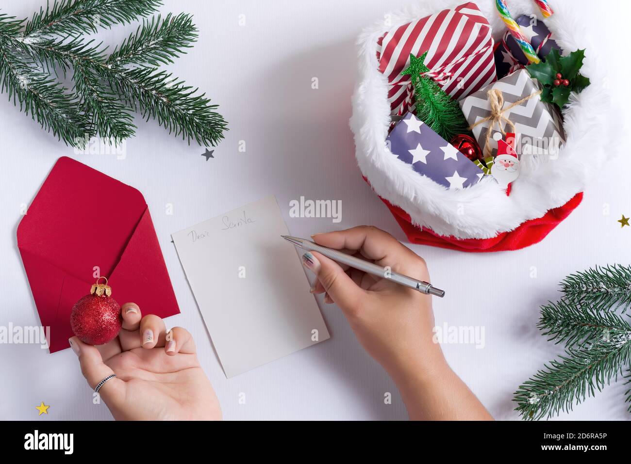 Carte de Noël avec les mains de la femme écrivant la lettre au Père Noël, tenant un réveil et un chapeau rouge rempli de différents cadeaux sur un fond gris clair, cop Banque D'Images