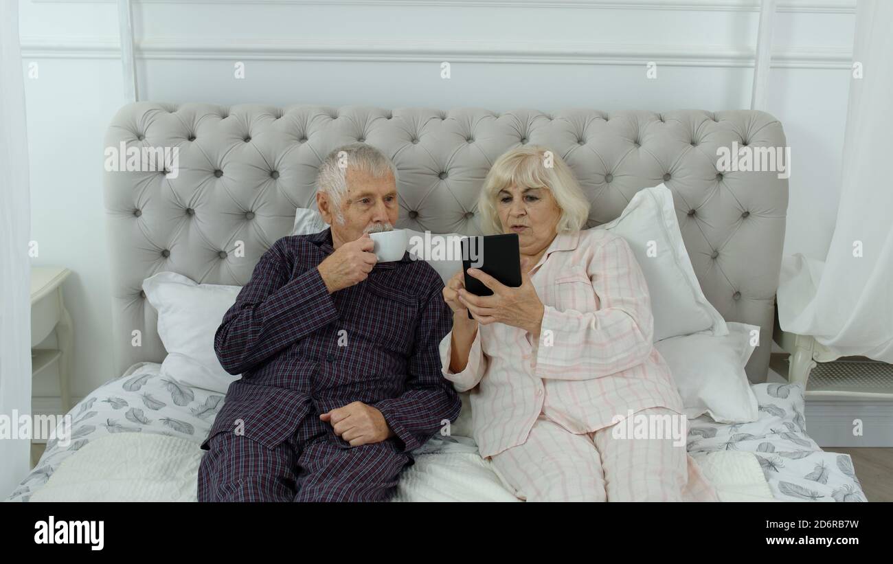 Un couple de personnes âgées portant un pyjama allongé sur un lit regardant  une tablette numérique rire et s'amuser. Grands-parents caucasiens matures,  mari et femme après le réveil faire des achats en