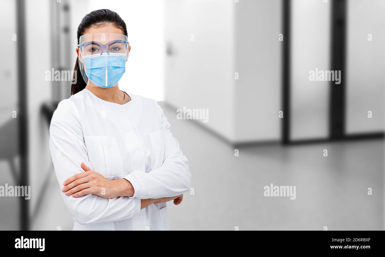 Portrait d'une femme médecin portant un écran de protection et un masque chirurgical, debout avec les bras croisés à l'hôpital de couloir. Banque D'Images