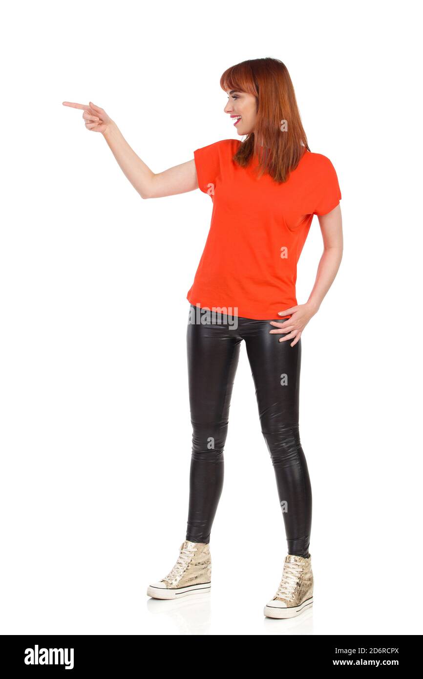Décontracté jeune femme en t-shirt orange, pantalon noir et baskets or est debout, pointant, parlant et regardant loin. Vue de profil. Studio complet Banque D'Images