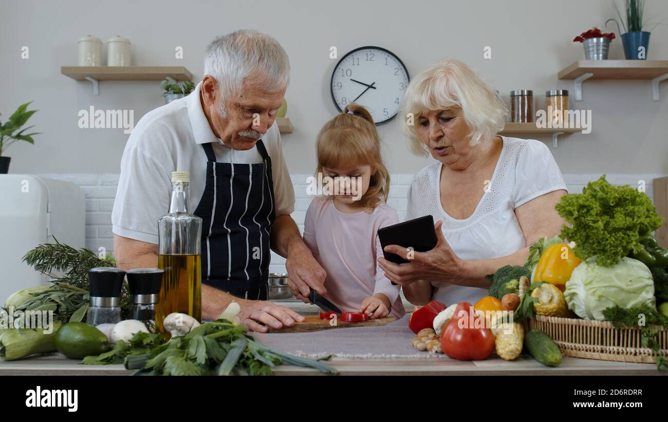 Les grands-parents âgés s'accouplent avec une petite-fille qui coupe des légumes pour la salade dans la cuisine. Homme et femme matures écoutant la recette, conseils de la tablette numérique. Alimentation saine. Régime alimentaire cru Banque D'Images