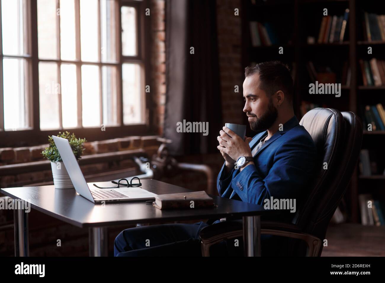 Savourez un café frais au travail. Un jeune homme plein de confiance dans un vêtement élégant et décontracté travaillant sur un ordinateur portable et buvant une boisson chaude tout en étant assis au travail Banque D'Images