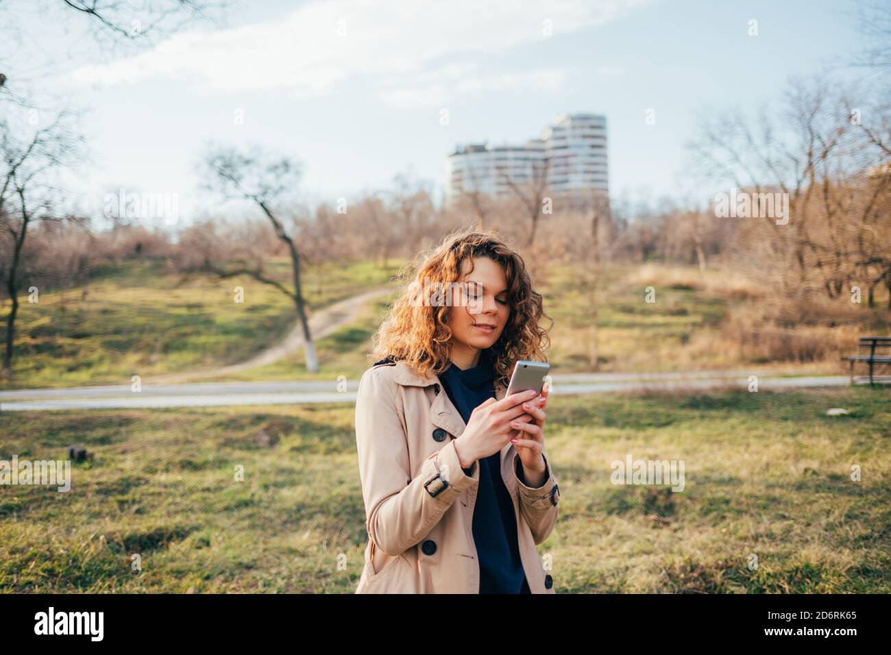 Jeune femme portant un manteau beige est debout à l'extérieur le jour de l'automne tenant des textos sur son smartphone. Banque D'Images
