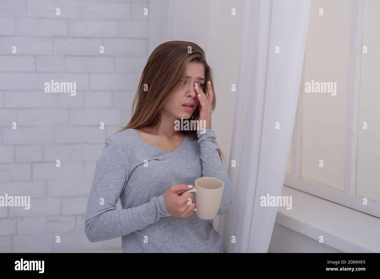 Une jeune femme malheureuse se tenant près de la fenêtre avec une tasse de café et pleurant, à l'intérieur Banque D'Images