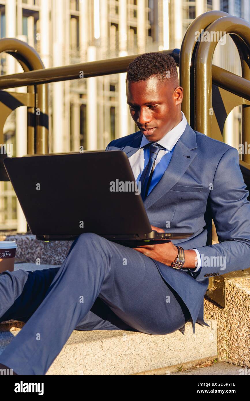 Vue du corps entier du gestionnaire financier afro-américain concentré dans élégant bleu costume formel assis sur les escaliers près de moderne bâtiment et lecture Banque D'Images