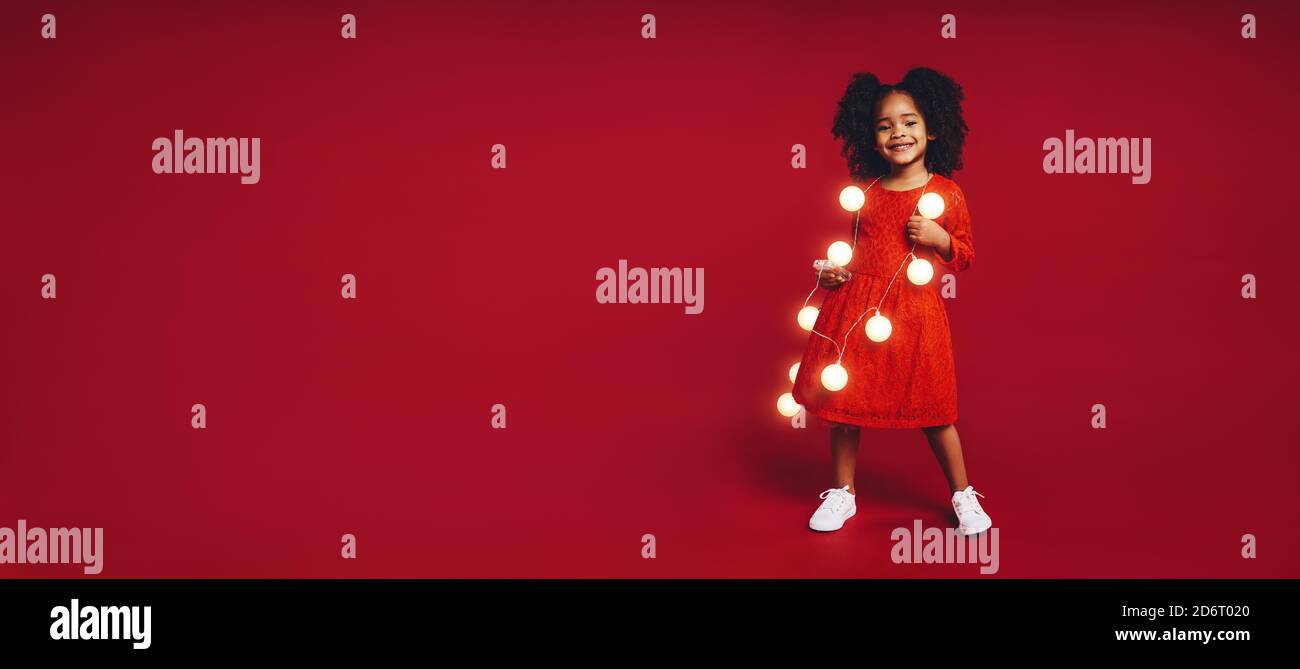 Souriante fille afro-américaine jouant avec des lumières de noël. Enfant debout sur fond rouge et portant des lumières de fées. Banque D'Images