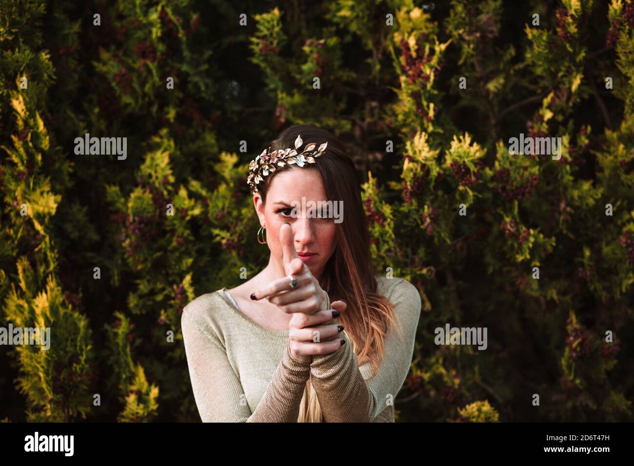 Jeune femme confiante portant une couronne à cheveux tendance contre l'abondance la verdure tout en pointant le doigt sur le pistolet et en regardant la caméra Banque D'Images