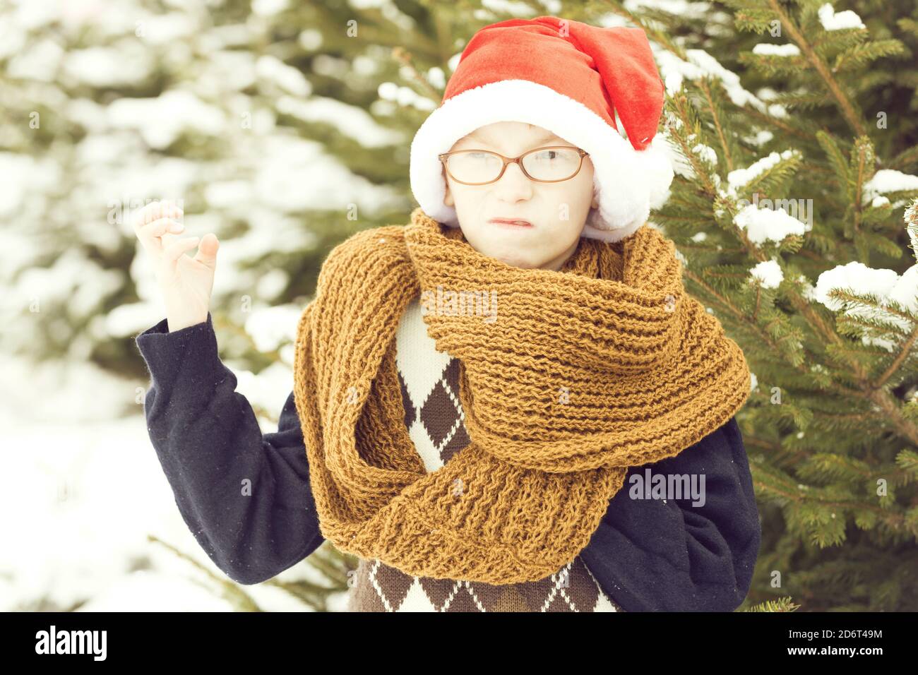 petit garçon en colère de noël ou petit enfant en lunettes, chandail, écharpe  tricotée tendance et chapeau de père noël rouge en hiver, en plein air sur  sapin vert avec neige sur