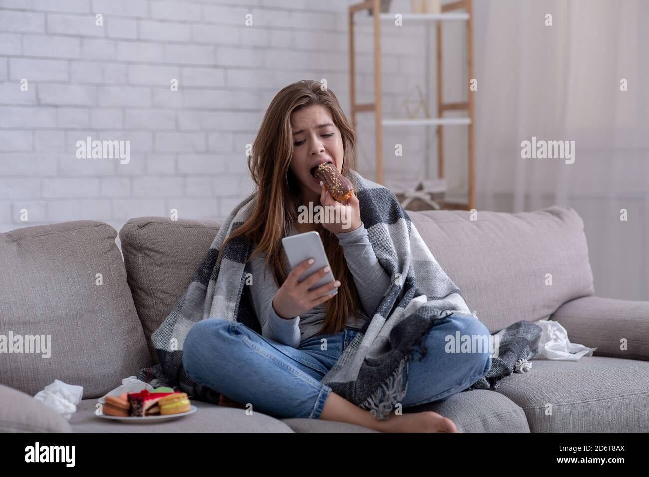 Concept d'alimentation émotionnelle. Énervé jeune femme avec une pâtisserie sucrée regardant le smartphone, en attendant un message important Banque D'Images