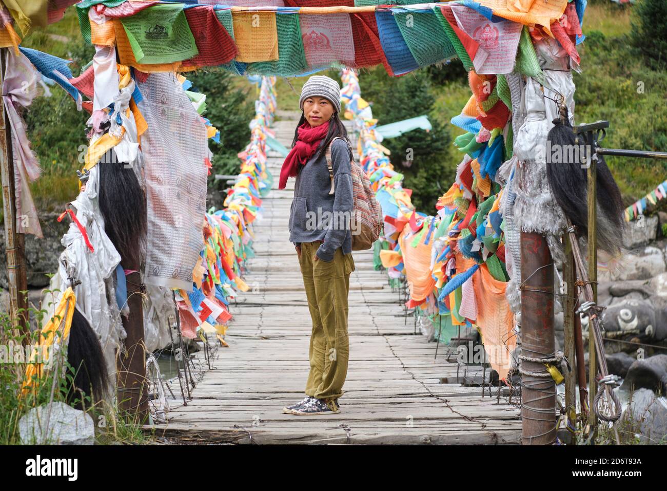 Jeune femme touristique ethnique dans des vêtements chauds et le chapeau regardant à la caméra tout en traversant un pont en bois simple à suspension étroite et longue décoration avec soupir Banque D'Images