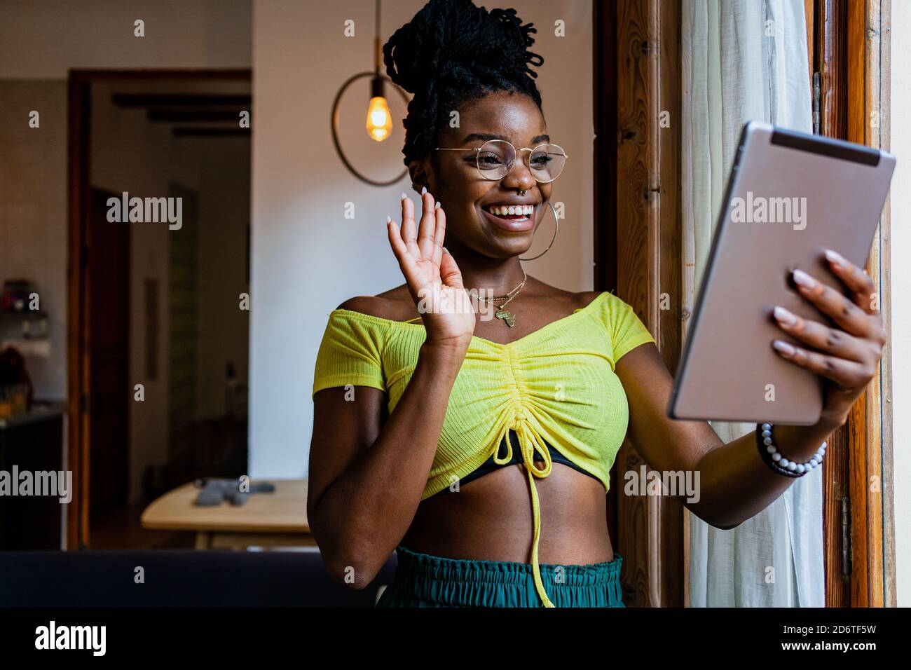Jeune femme noire gaie en crop top et lunettes de vue en agitant main et sourire tout en discutant vidéo sur tablette à accueil Banque D'Images