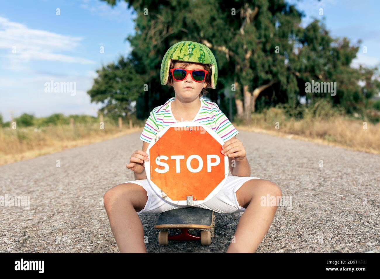 Enfant élégant en melon d'eau décoratif tête de port et lunettes de soleil  assis Sur le skateboard tout en affichant un panneau avec UN TITRE D'ARRÊT  sur la route Photo Stock -