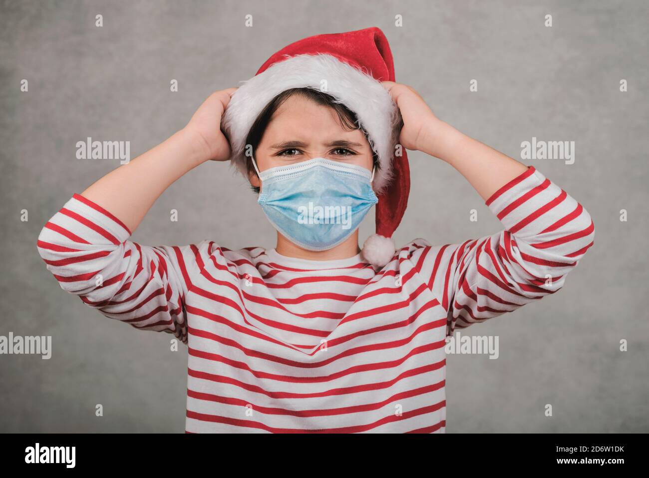 Joyeux Noël, un enfant en colère avec un masque médical portant le chapeau du Père Noël avec ses mains sur la tête Banque D'Images