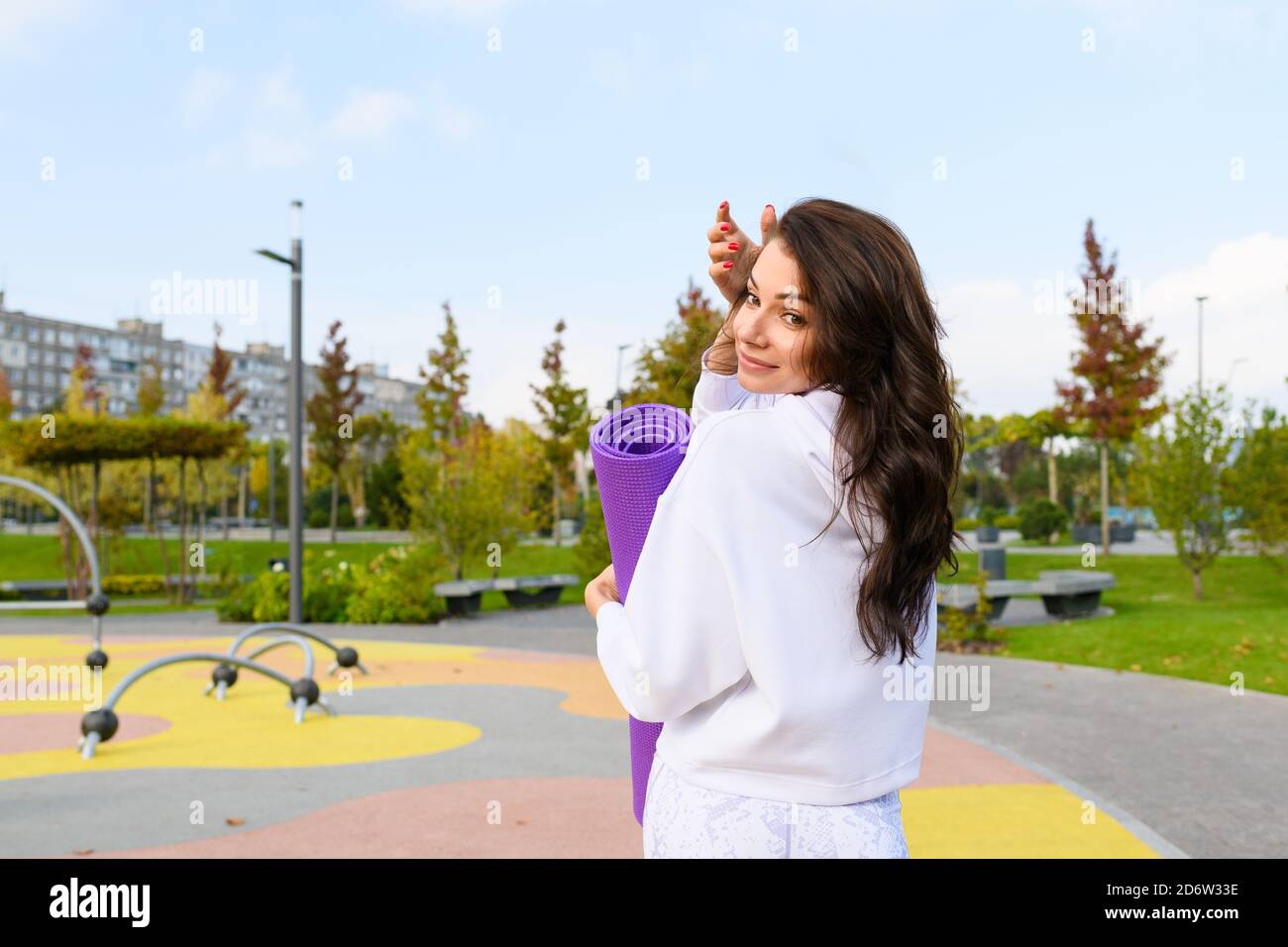 Sportive brunette femme à capuche blanche tenir le tapis violet à l'aire de jeux du parc de la ville, l'entraînement, la forme physique, s'étirant à l'extérieur Banque D'Images
