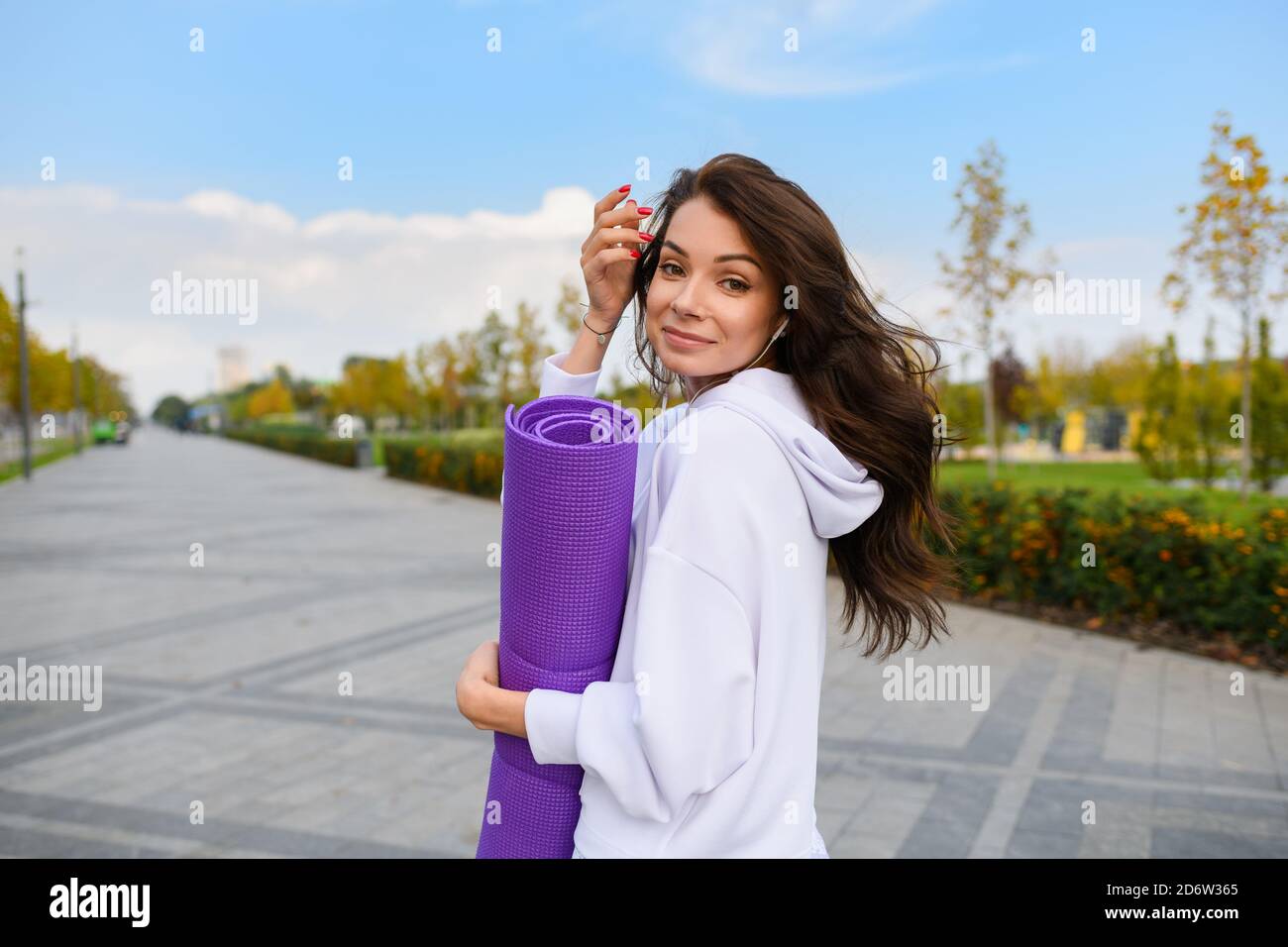 Brunette jeune femme en blanc vêtements de sport avec écouteurs tient violet et en regardant en arrière-plan le parc de la ville Banque D'Images