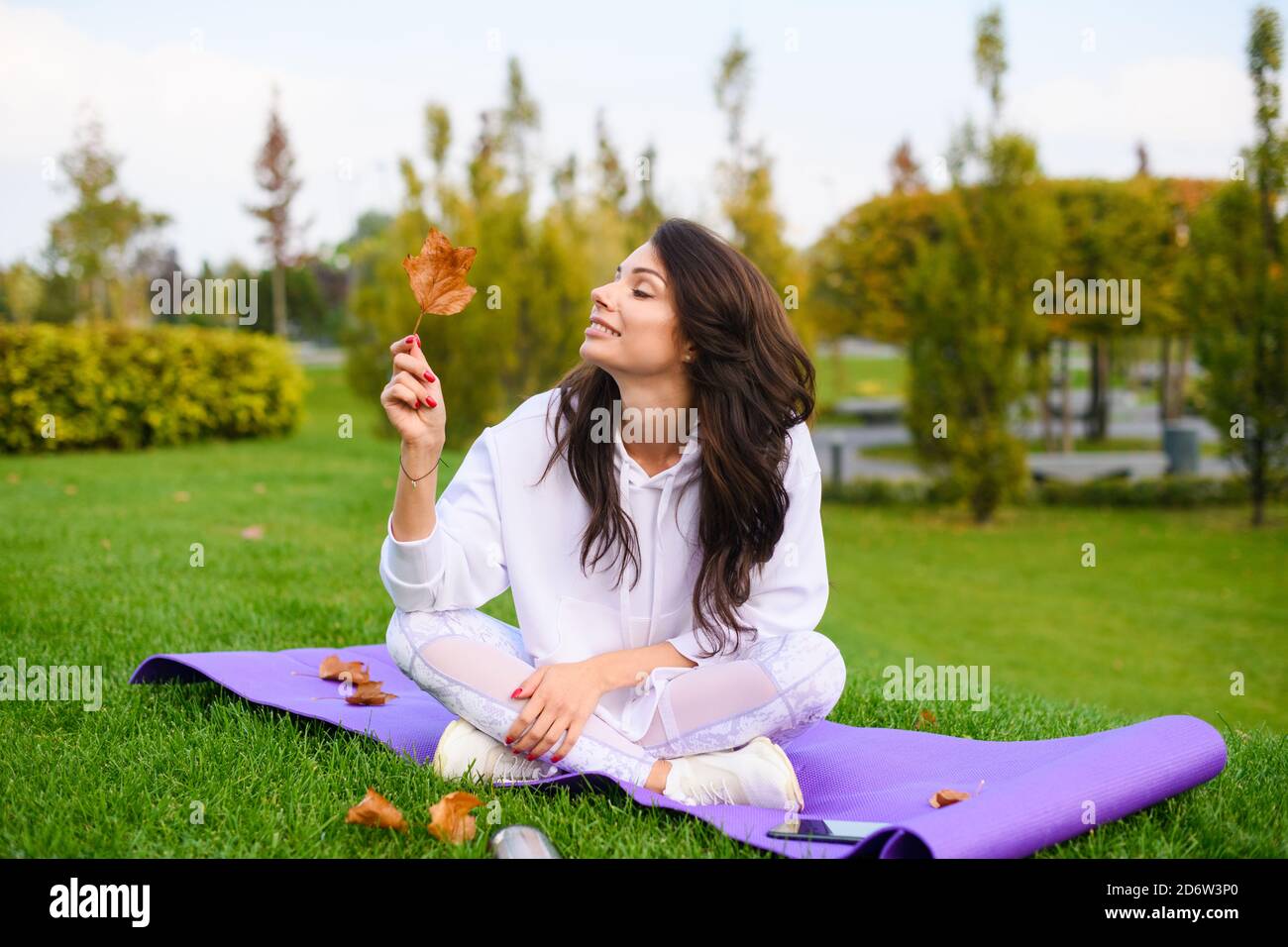 Belle jeune femme s'assoit au tapis de sport violet dans le parc d'automne de la ville, prendre et regarder une feuille sèche après l'entraînement en plein air Banque D'Images
